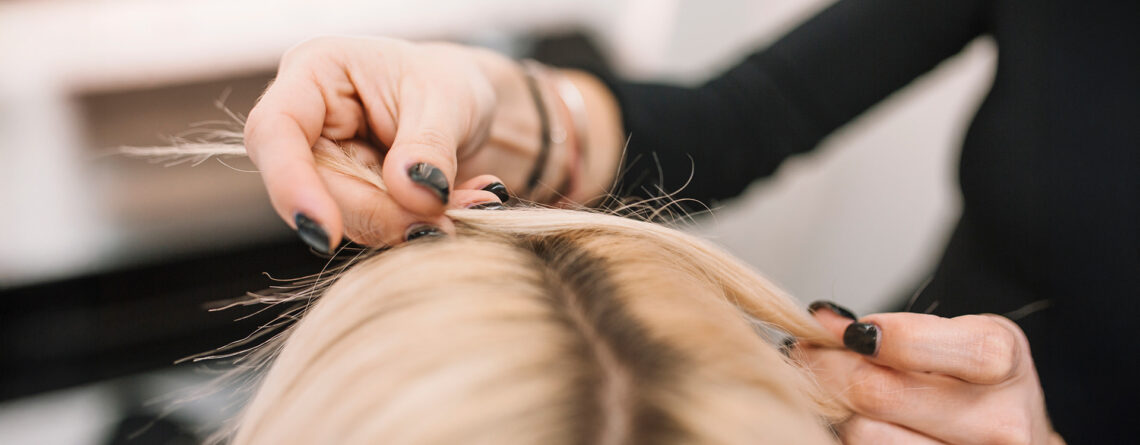 Zagęszczanie włosów w salonie fryzjerskim – stylistka pracuje przy przedziałku na głowie klientki, unosząc pasma cienkich, blond włosów.