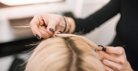 Zagęszczanie włosów w salonie fryzjerskim – stylistka pracuje przy przedziałku na głowie klientki, unosząc pasma cienkich, blond włosów.